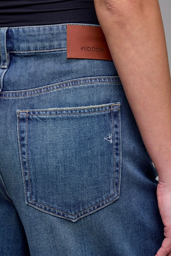 Close-up of blue jeans with a brown leather patch and 'Hidden' branding.