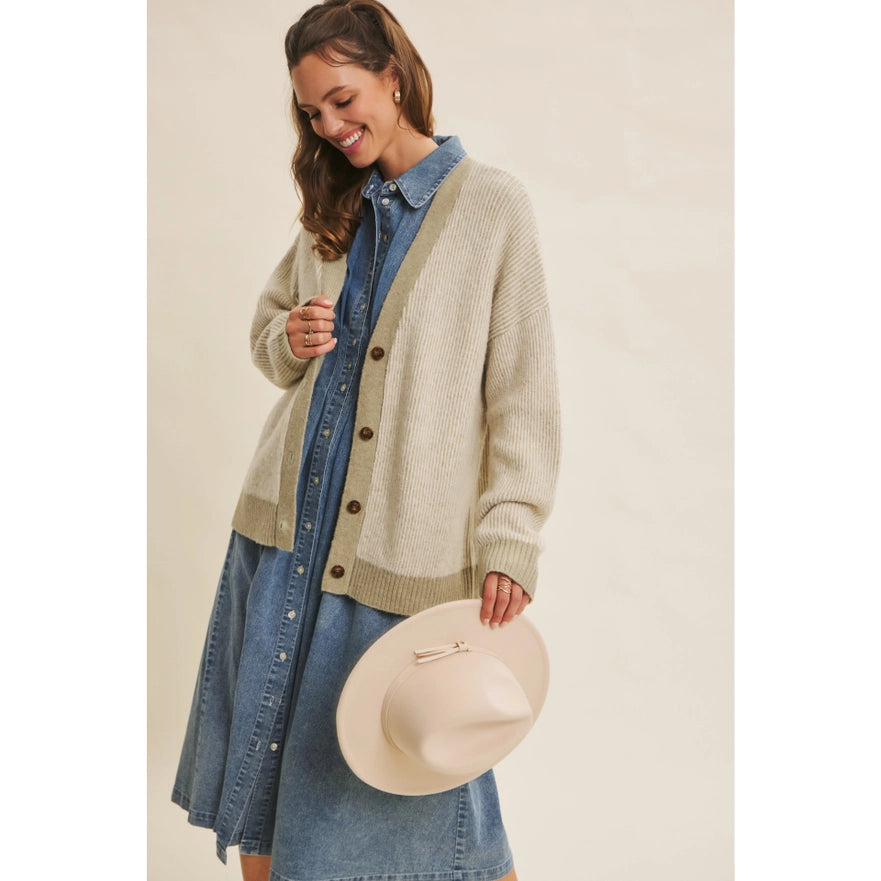 Woman wearing a beige cardigan over a denim dress, holding a beige hat against a neutral background