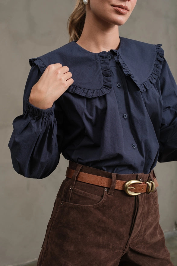Person wearing a navy blue blouse with ruffled collar and brown pants against a neutral background