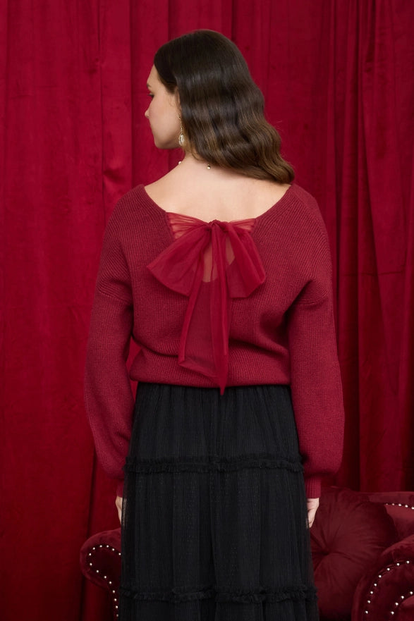 Woman wearing a red sweater with a bow detail in front of a red curtain.