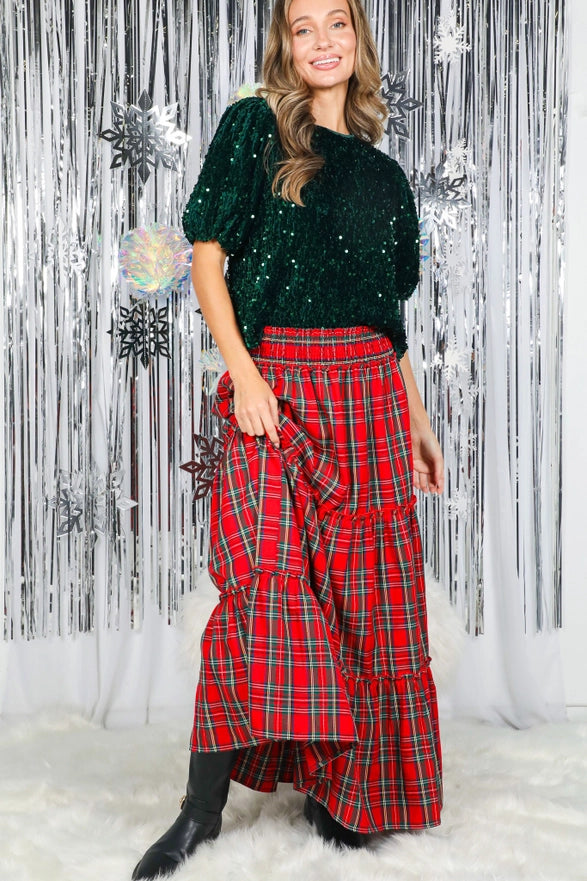 Woman wearing a green sequin top and red plaid skirt against a decorative silver and white background.