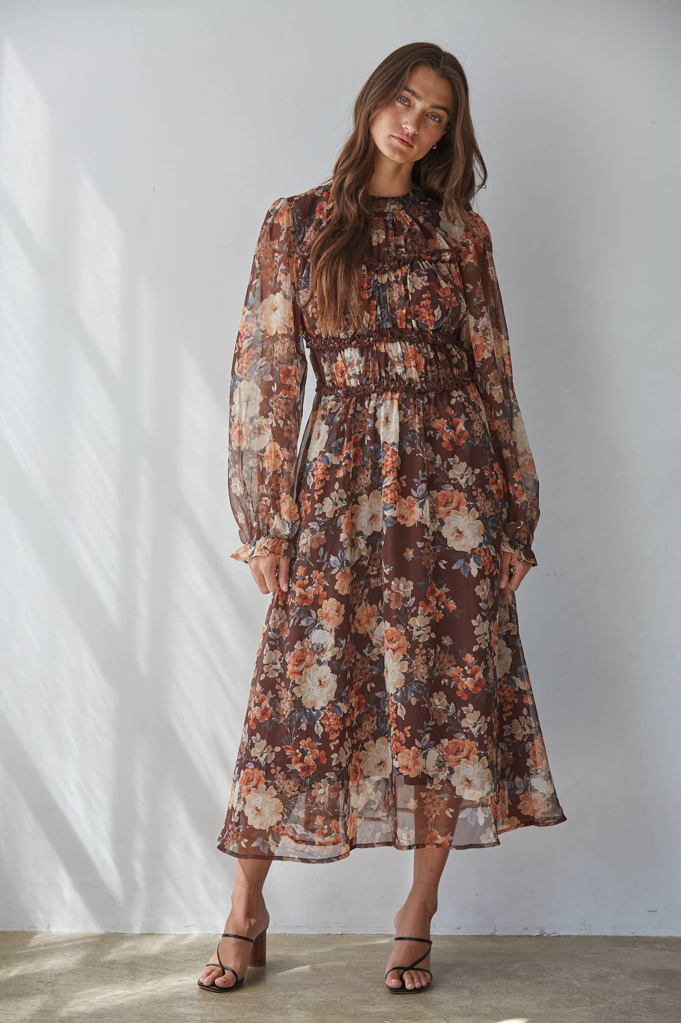 A woman modeling a long-sleeve midi dress with a dense floral print in earthy tones of brown, rust, beige, and muted orange, standing against a white wall with soft shadow patterns.