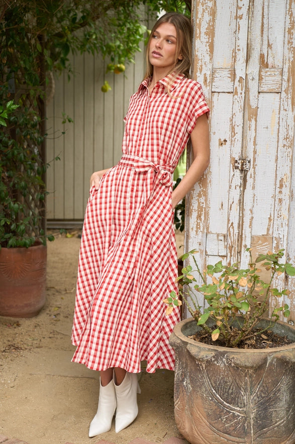 Woman in a red and white checkered dress standing in an outdoor setting with plants and a wooden wall.