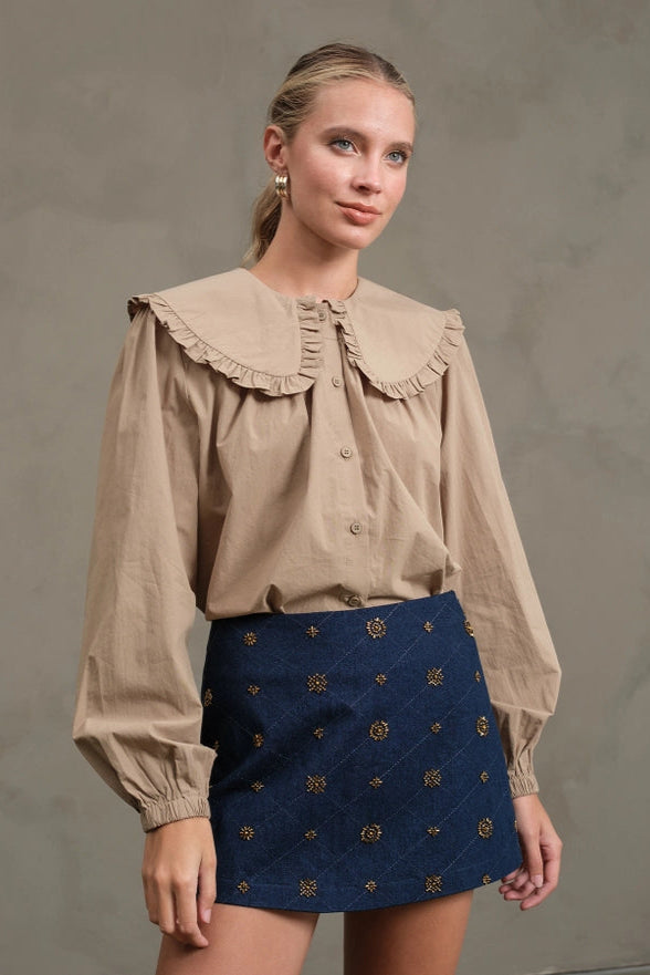 Woman wearing a beige blouse with a ruffled collar and a navy skirt with gold patterns against a gray background
