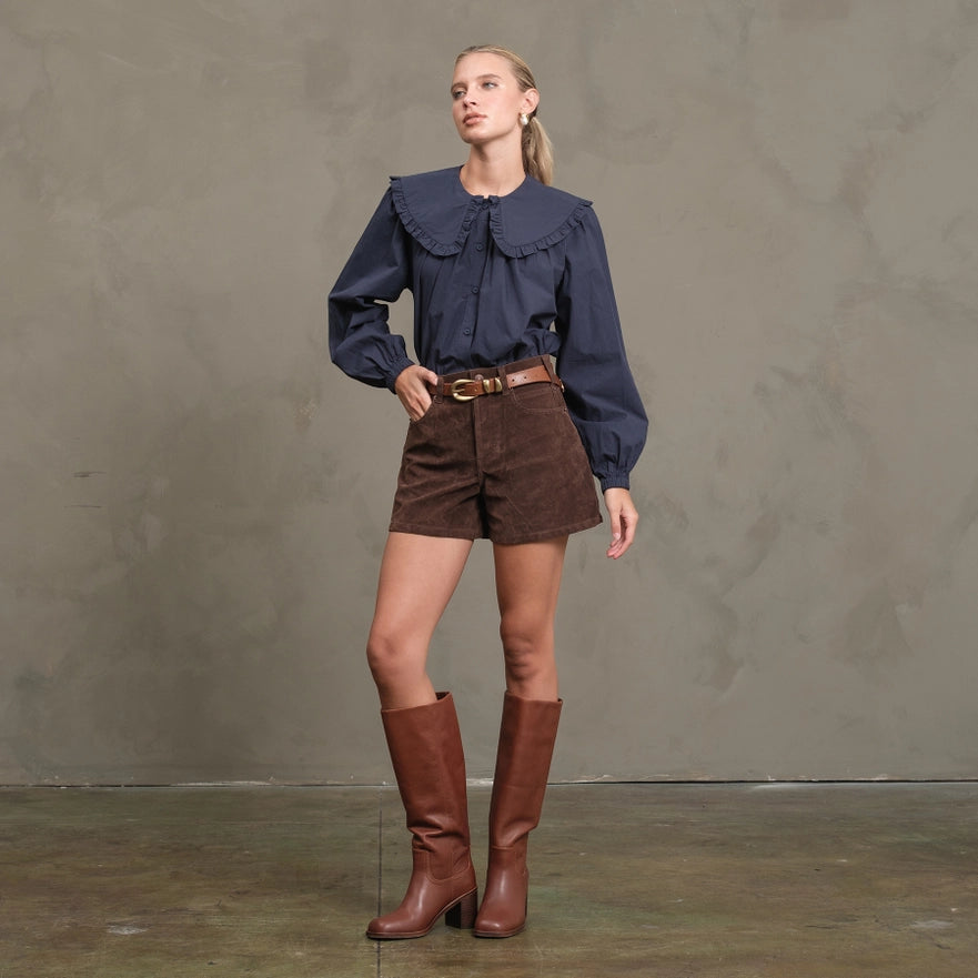 Woman wearing a blue blouse, brown shorts, and brown knee-high boots against a gray background