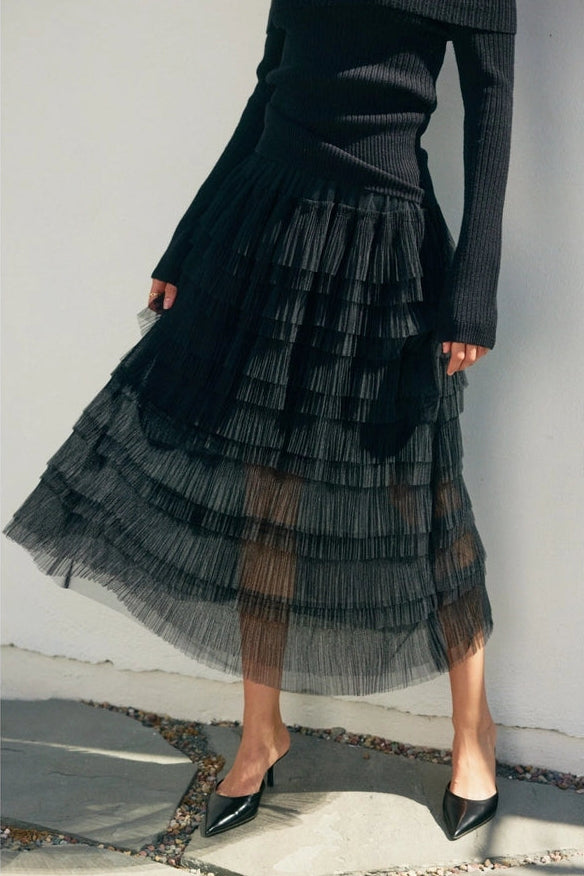 Person wearing a black ruffled skirt and black sweater against a white wall.