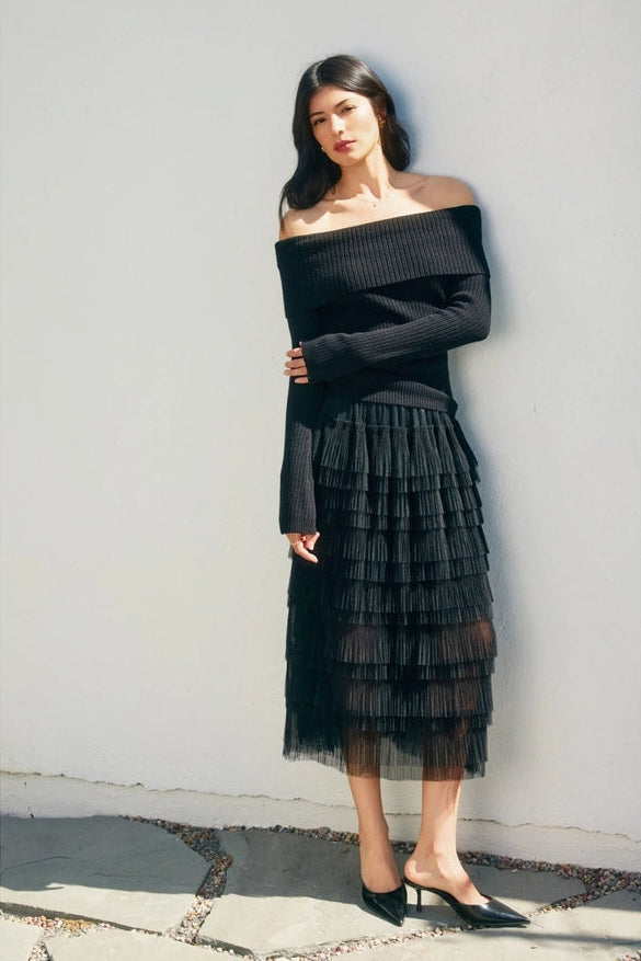 Woman wearing a black off-shoulder top and tiered skirt against a white wall.