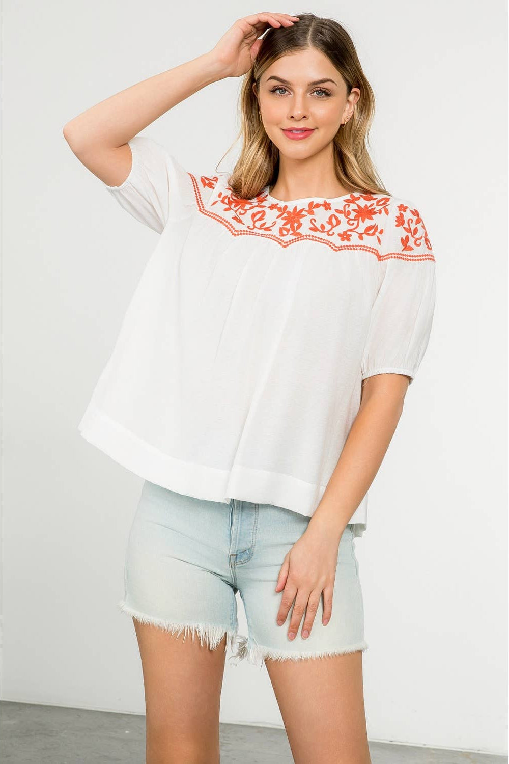 Woman wearing a white blouse with red embroidery and denim shorts on a plain background