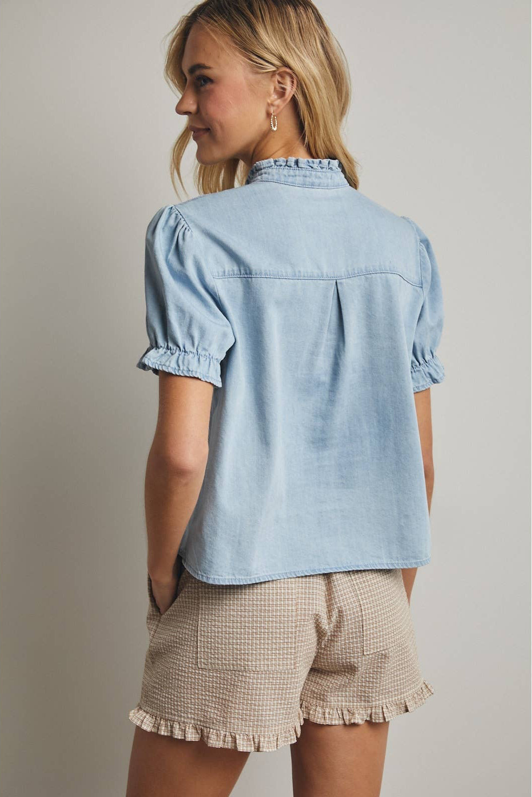Woman wearing a light blue denim shirt and beige shorts against a neutral background