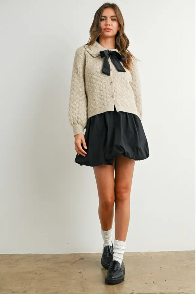 Woman wearing a beige knitted sweater with a black bow and a black skirt on a white background