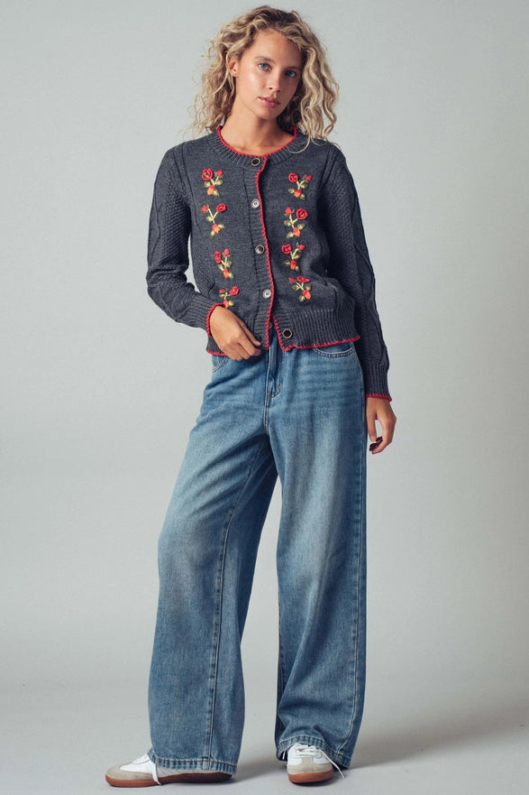 Woman wearing a dark denim jacket with floral embroidery and blue jeans on a gray background