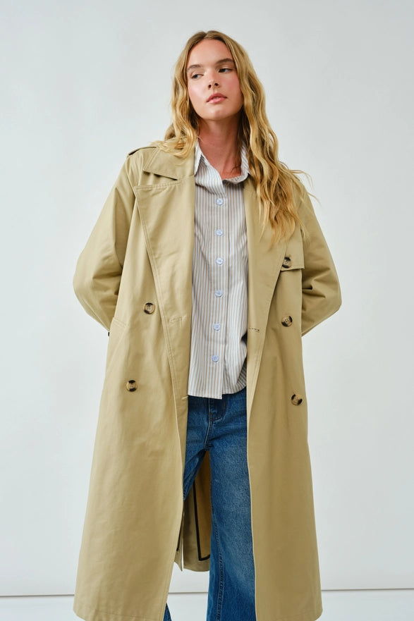 Woman wearing a beige trench coat over a striped shirt and jeans on a white background