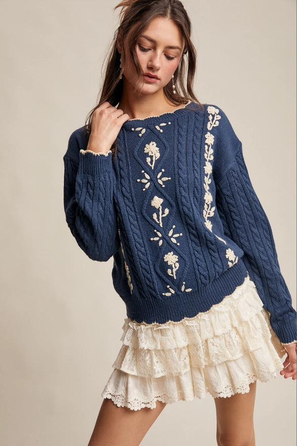 Woman wearing a blue sweater with floral patterns and a white skirt on a beige background