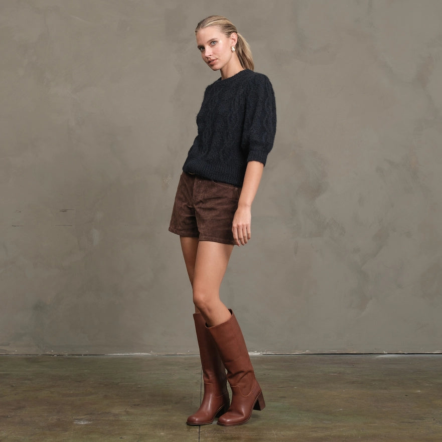 Woman wearing a dark sweater, brown shorts, and brown knee-high boots against a gray background