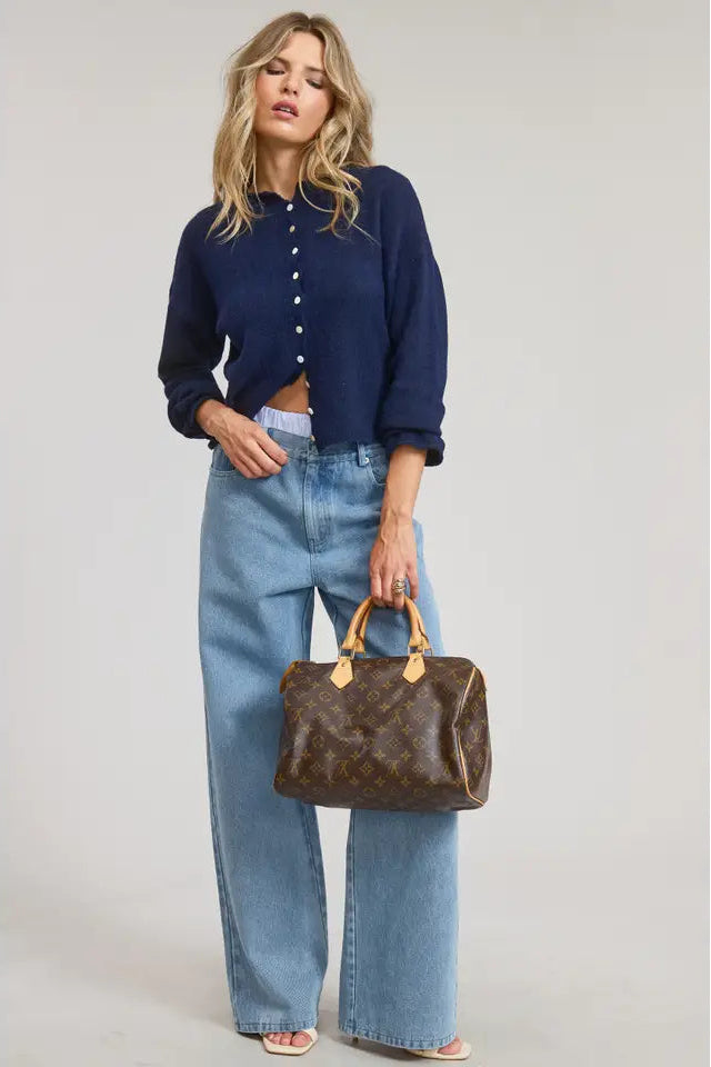 Woman wearing a navy blue shirt and light blue jeans, holding a brown handbag on a plain background