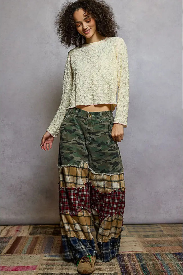 Person wearing a cream lace top and patchwork pants against a gray wall.