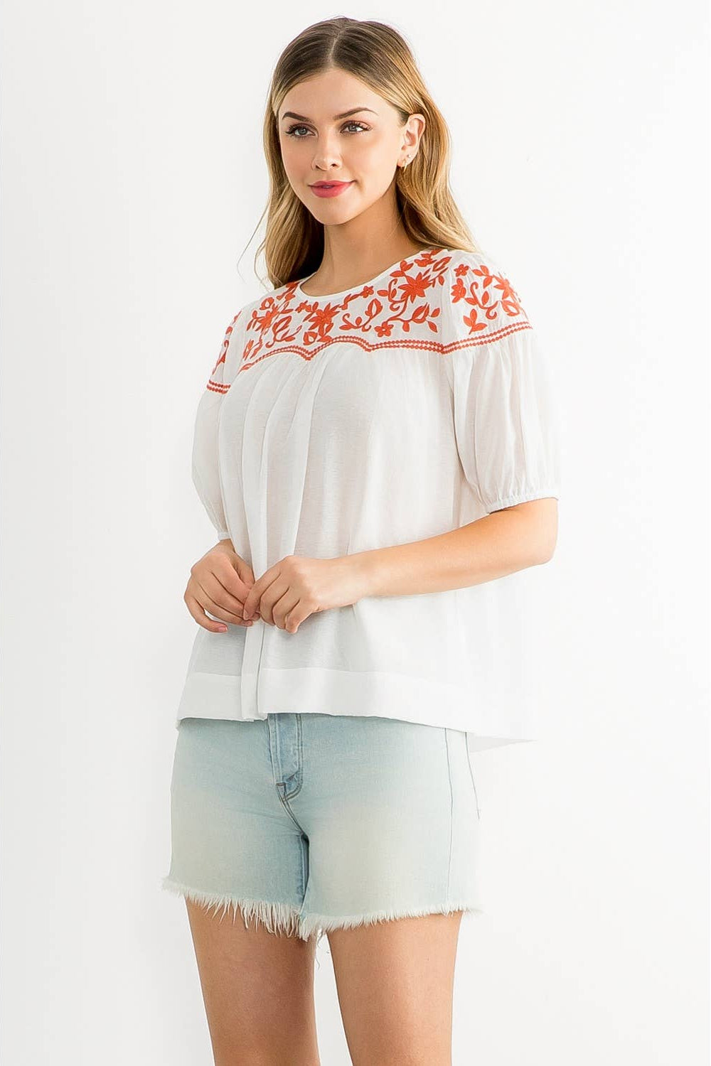 Woman wearing a white blouse with red embroidery and denim shorts on a white background