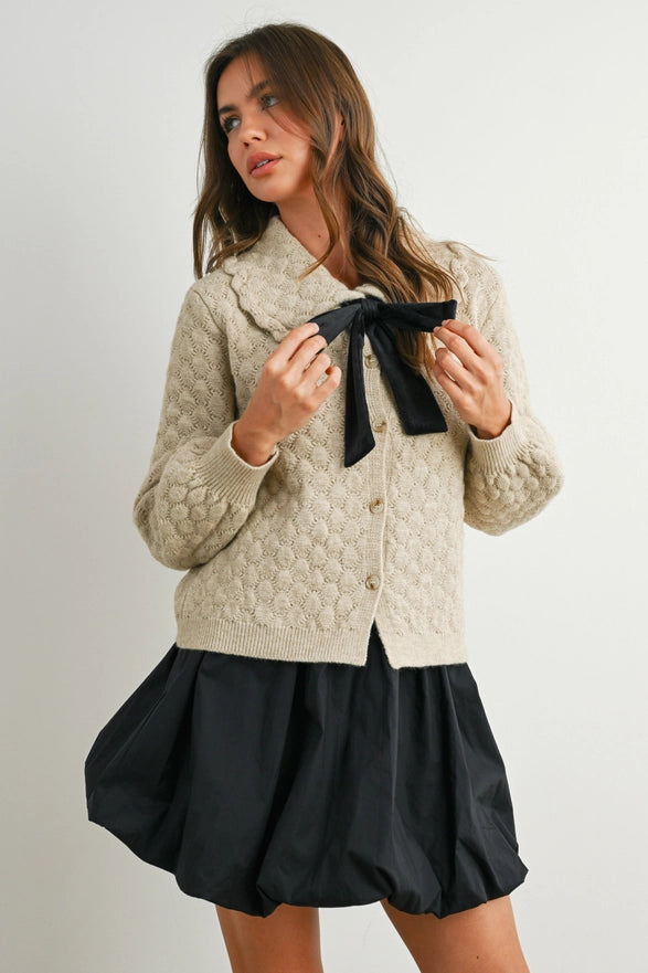 Woman wearing a textured beige cardigan with a black bow and a black skirt on a plain background
