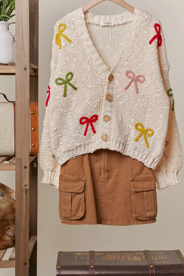 Knitted cardigan with colorful bow designs and brown shorts on a hanger against a neutral wall.