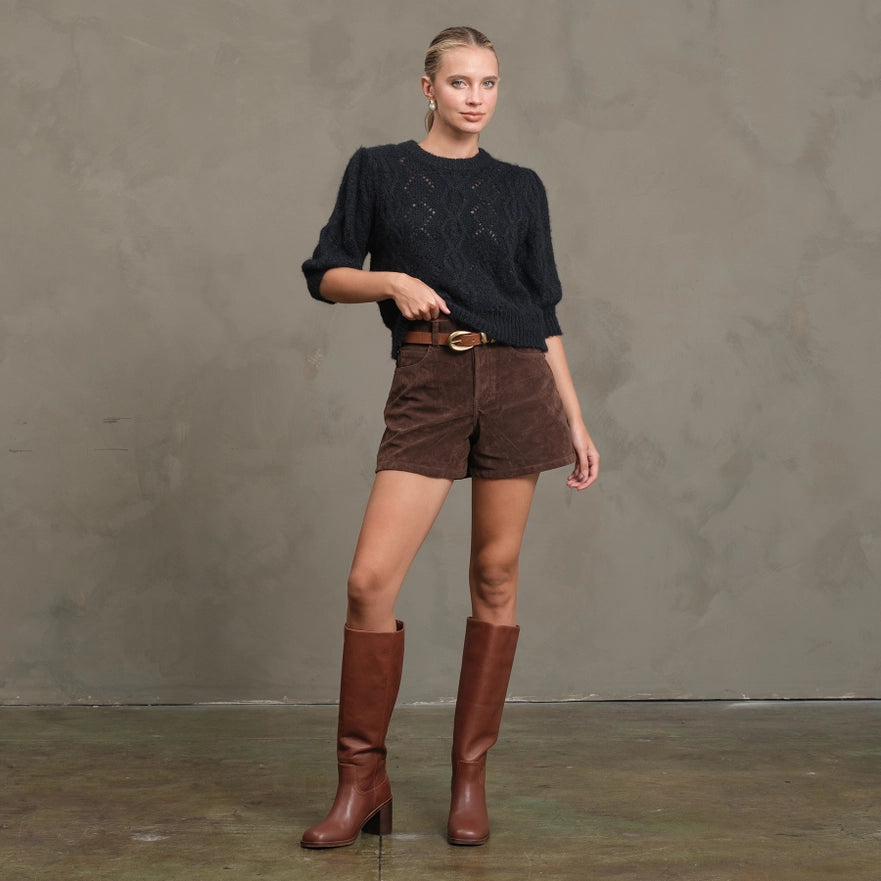 Woman wearing a black sweater and brown shorts with brown boots against a gray background