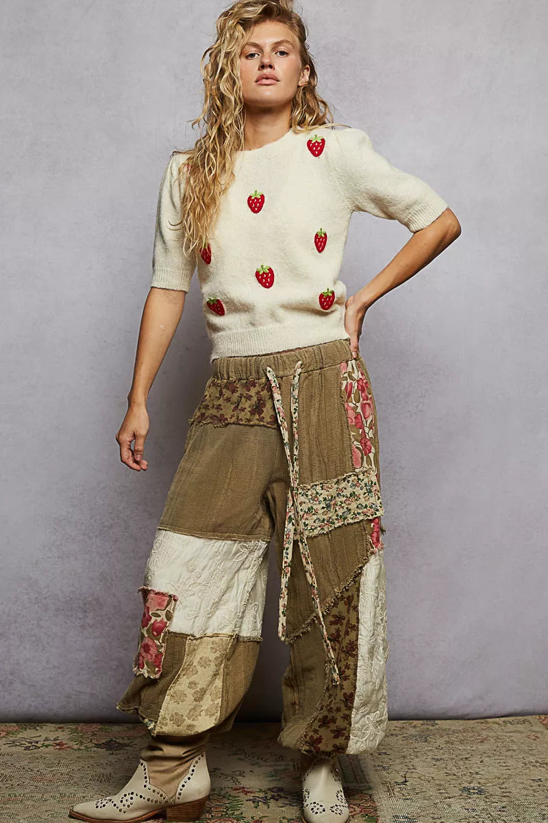 Woman wearing a cream sweater with red strawberry pattern and patchwork pants against a gray background