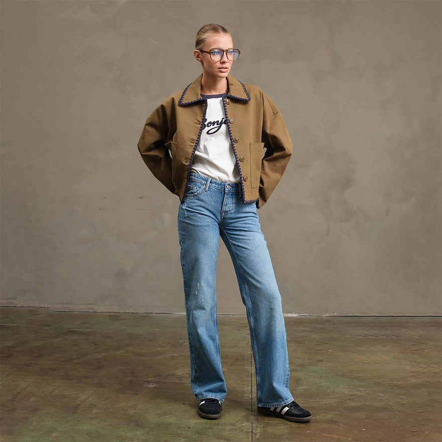 Person wearing a brown jacket, white shirt with text, and blue jeans against a plain background