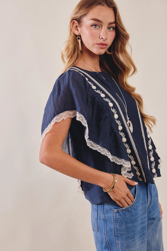 Woman wearing a navy blue top with white lace details and blue jeans on a plain background