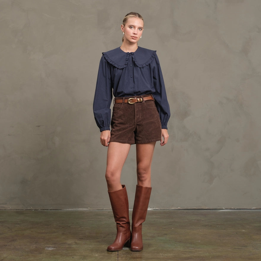 Woman wearing a blue blouse, brown shorts, and brown boots against a gray background