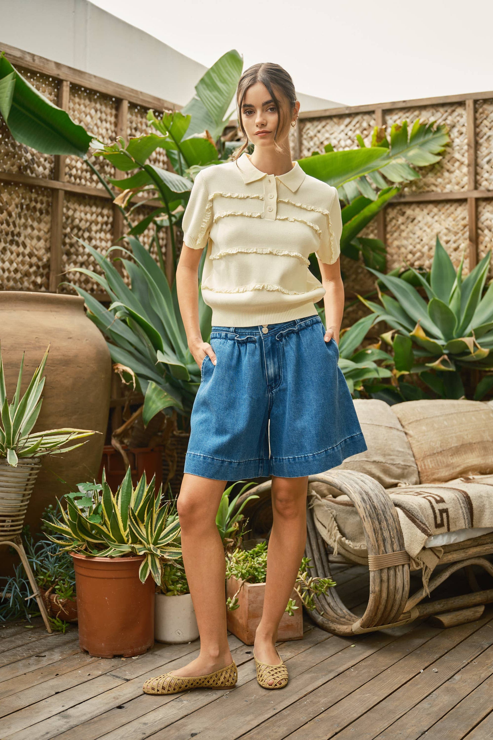 Woman in a yellow top and blue denim skirt standing in a garden setting with plants and furniture.