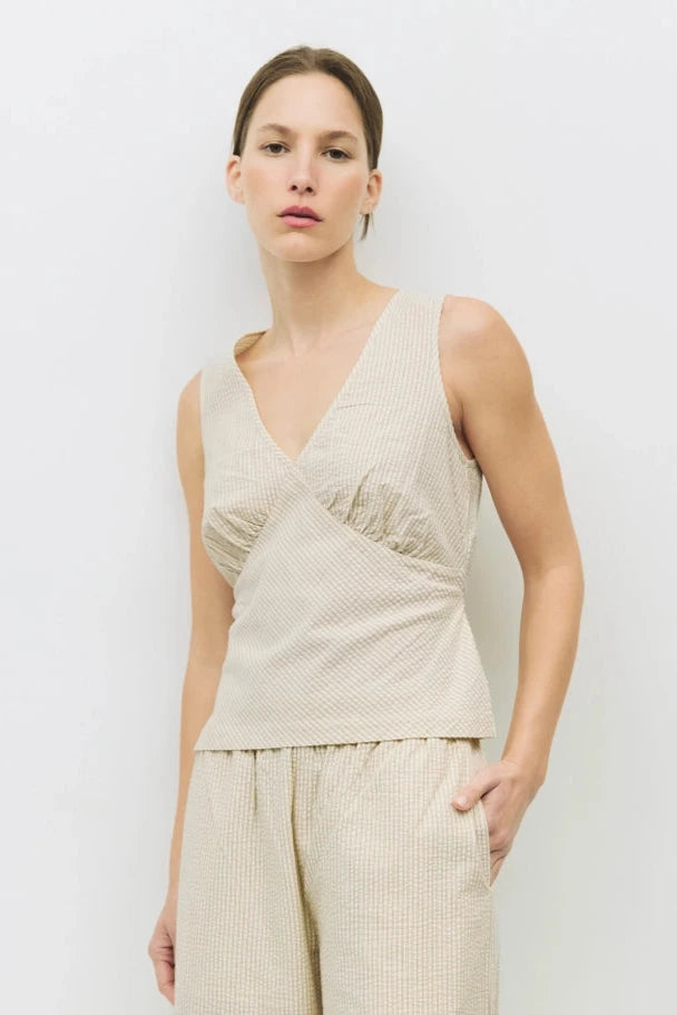 A female model wearing a beige sleeveless seersucker top with a V-neckline, bust seam shirring, and side ties, paired with matching pants.