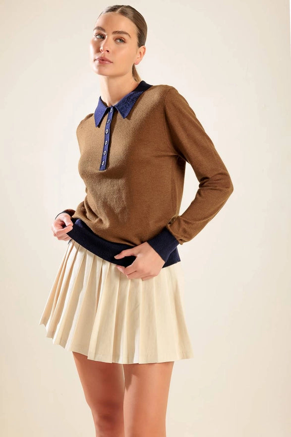 Woman wearing a brown sweater with a blue shirt underneath and a beige pleated skirt on a plain background