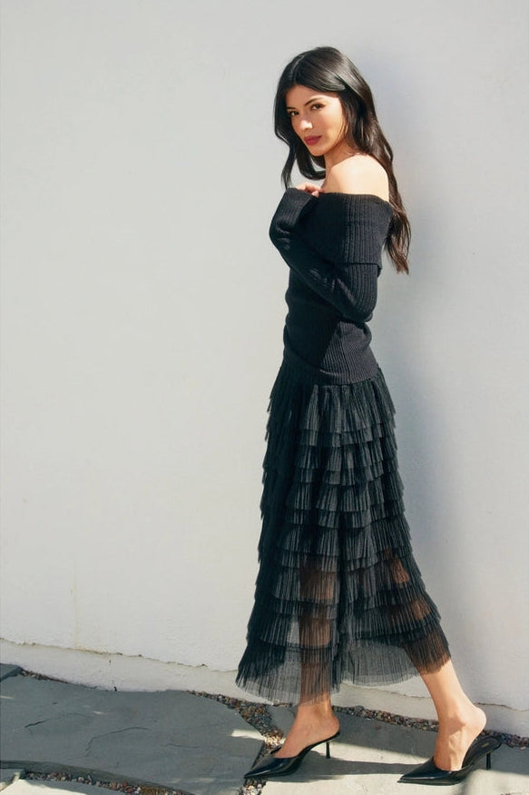 Woman wearing a black off-shoulder top and tiered skirt against a white wall.