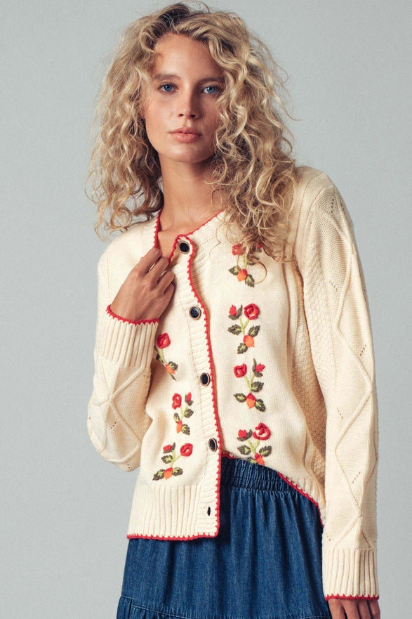 Woman wearing a cream cardigan with floral embroidery on a gray background