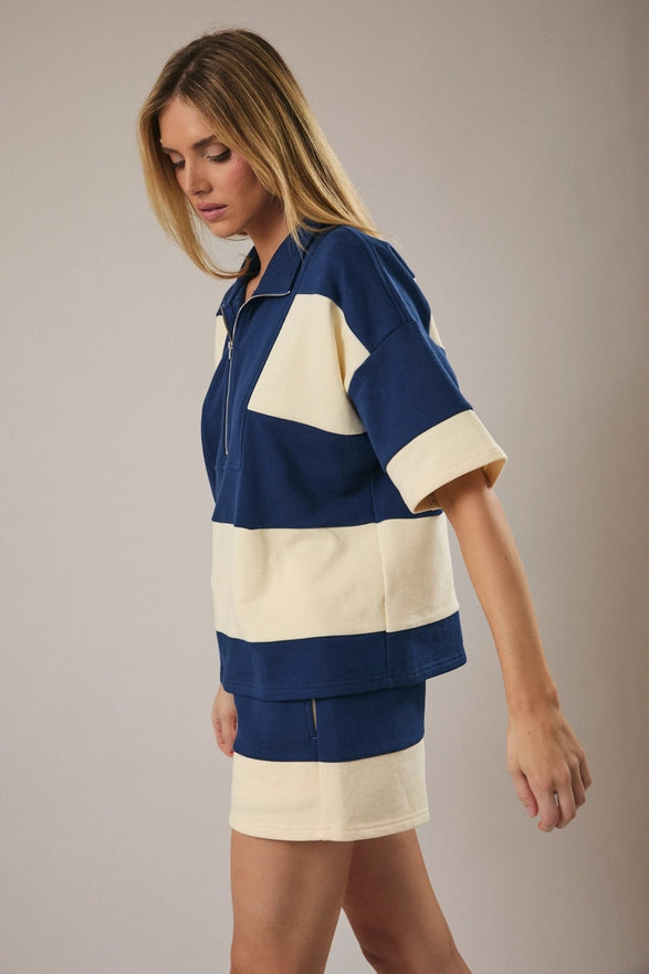 Model is wearing a matching cream and navy striped set with shorts and a half zip polo like top.