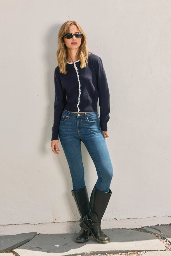 Woman wearing a navy sweater, blue jeans, and black boots against a light gray wall.