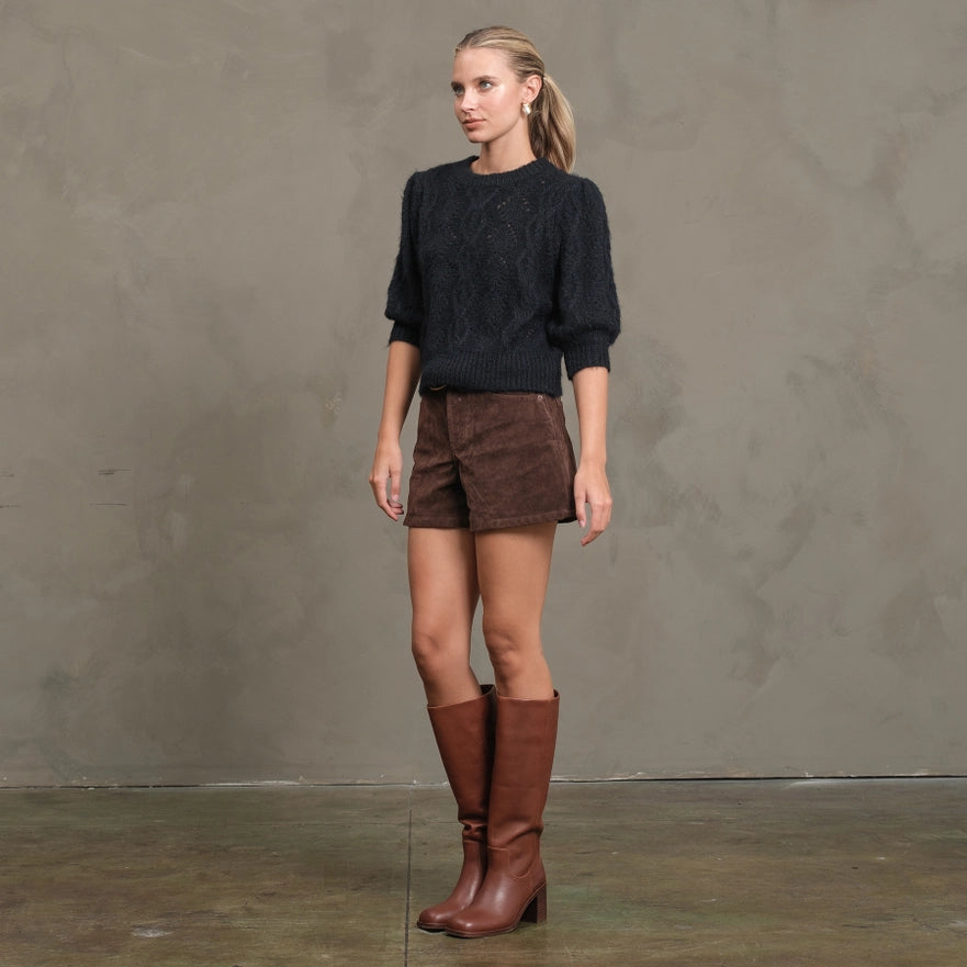 Woman wearing a dark sweater, brown shorts, and brown knee-high boots against a plain background