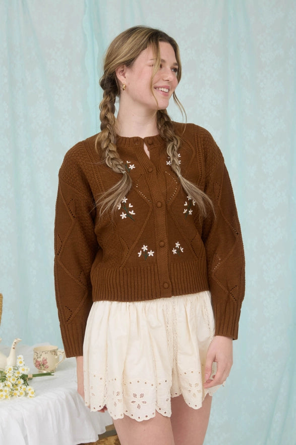 Woman wearing a brown cardigan with star patterns and a white lace skirt against a light blue curtain background.