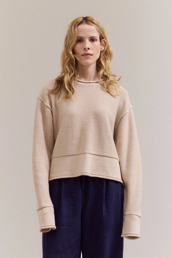 Woman wearing a beige sweater and dark pants against a plain background