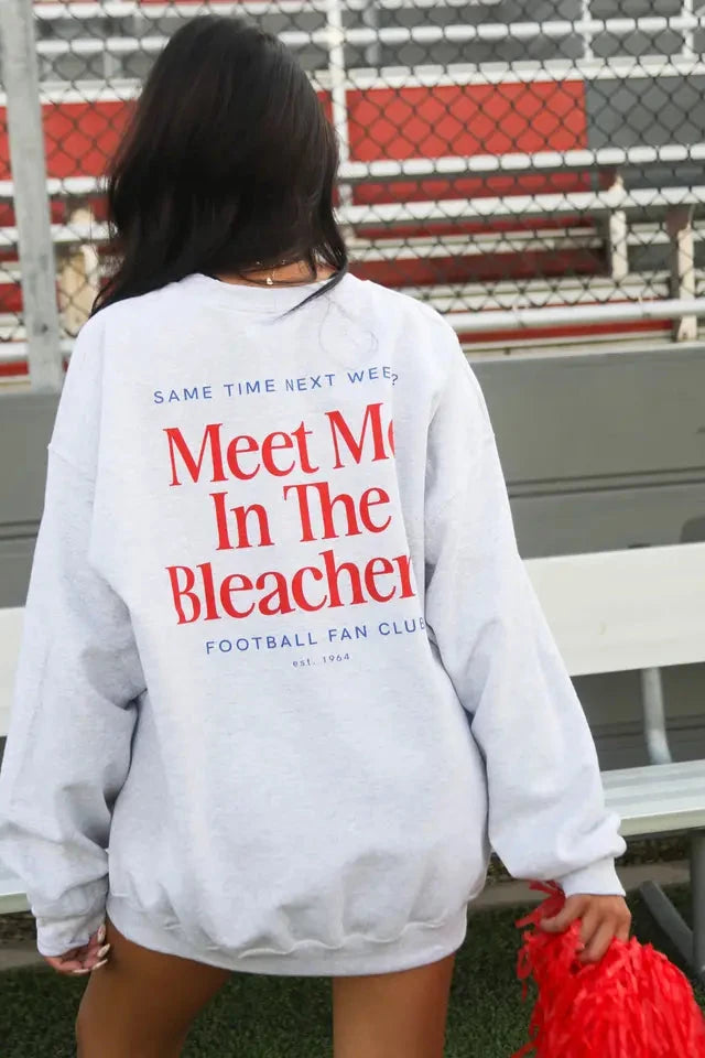 Person wearing a sweatshirt with 'Meet Me In The Bleachers' text at a sports event.
