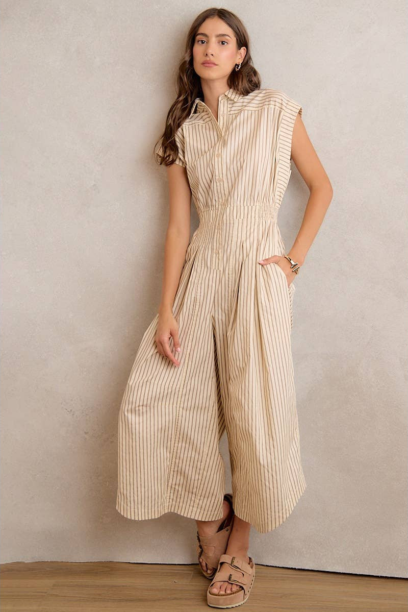 Woman wearing a beige and white striped jumpsuit against a plain wall.