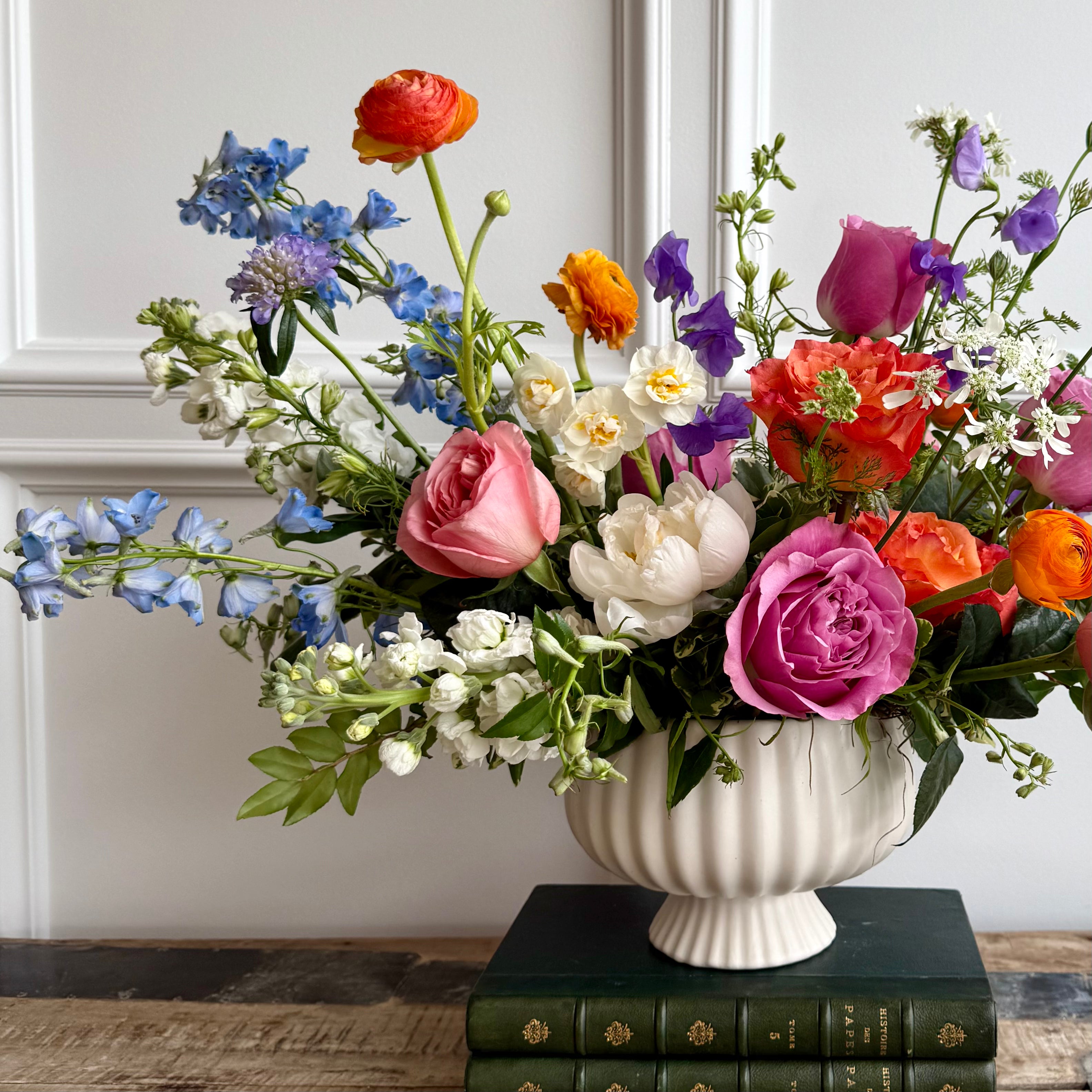 A vibrant floral arrangement in a white ceramic vase featuring pink, orange, and purple blooms including roses, ranunculus, sweet peas, and peonies, with touches of white and blue flowers. The bouquet is displayed on stacked green vintage books atop a rustic wooden table, with a white panel wall in the background.
