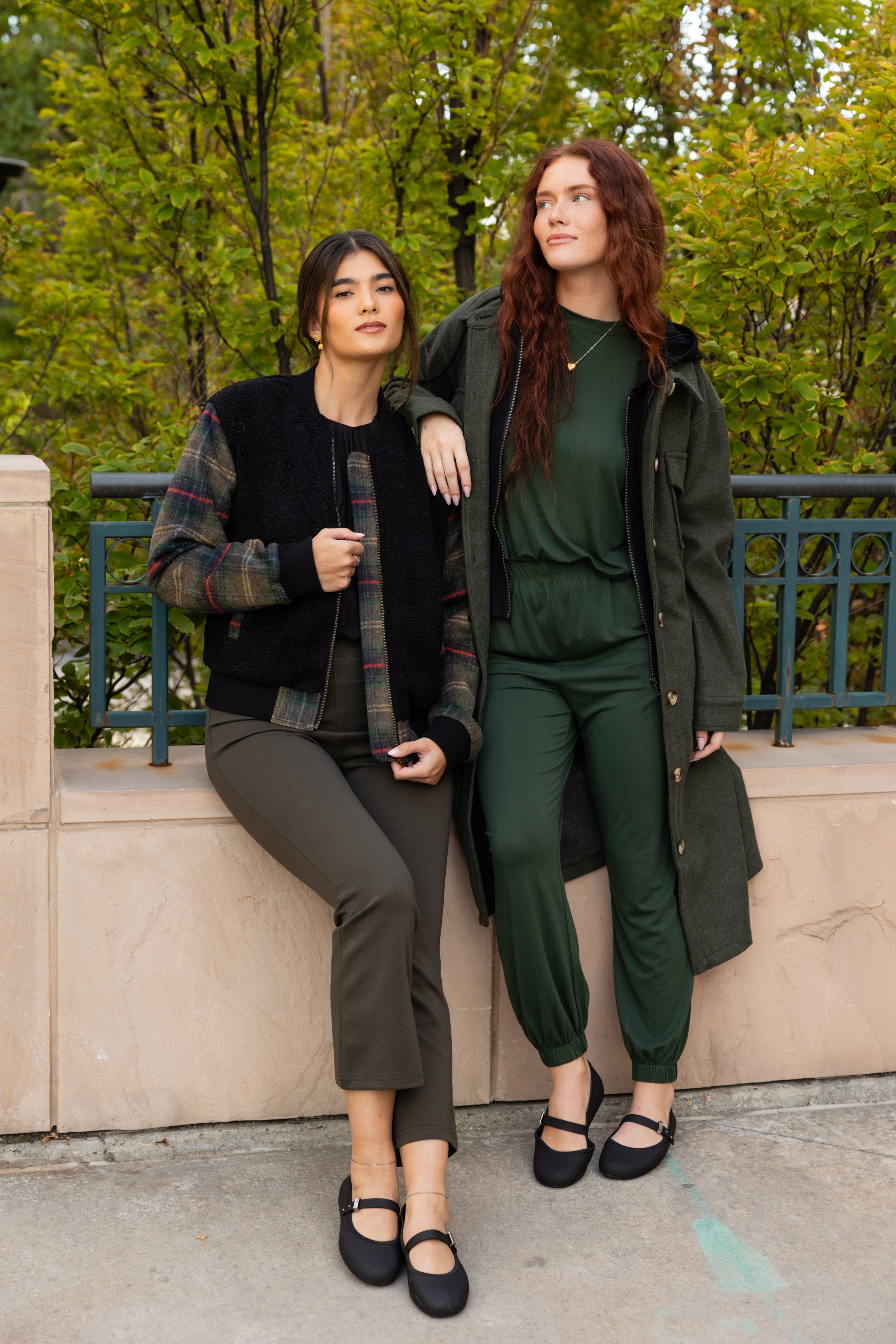 A woman wearing a black fleece bomber jacket with plaid sleeves in green, red, and neutral colors, paired with dark pants, standing by a greenery backdrop.