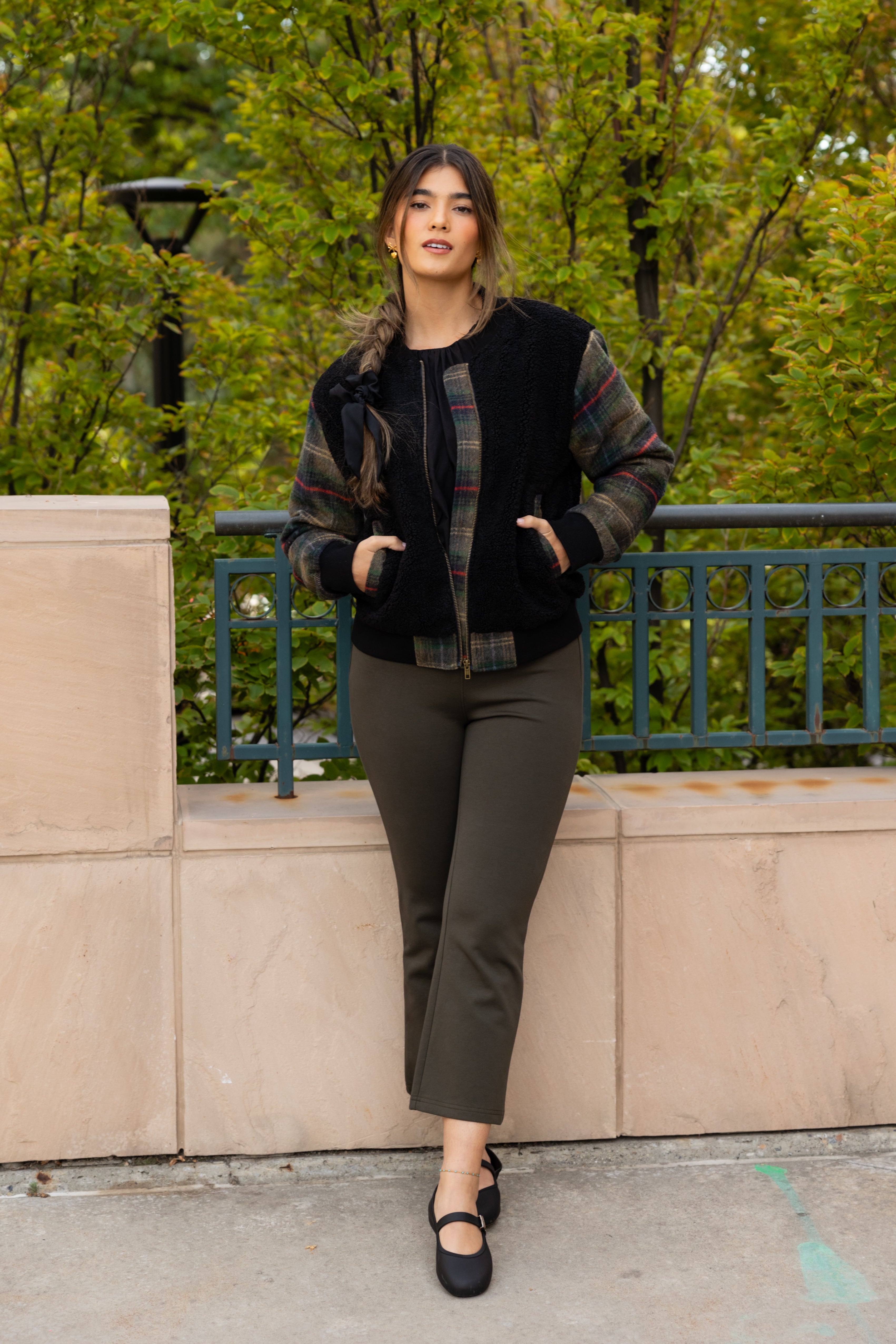 A woman wearing a black fleece bomber jacket with plaid sleeves in green, red, and neutral colors, paired with dark pants, standing by a greenery backdrop.