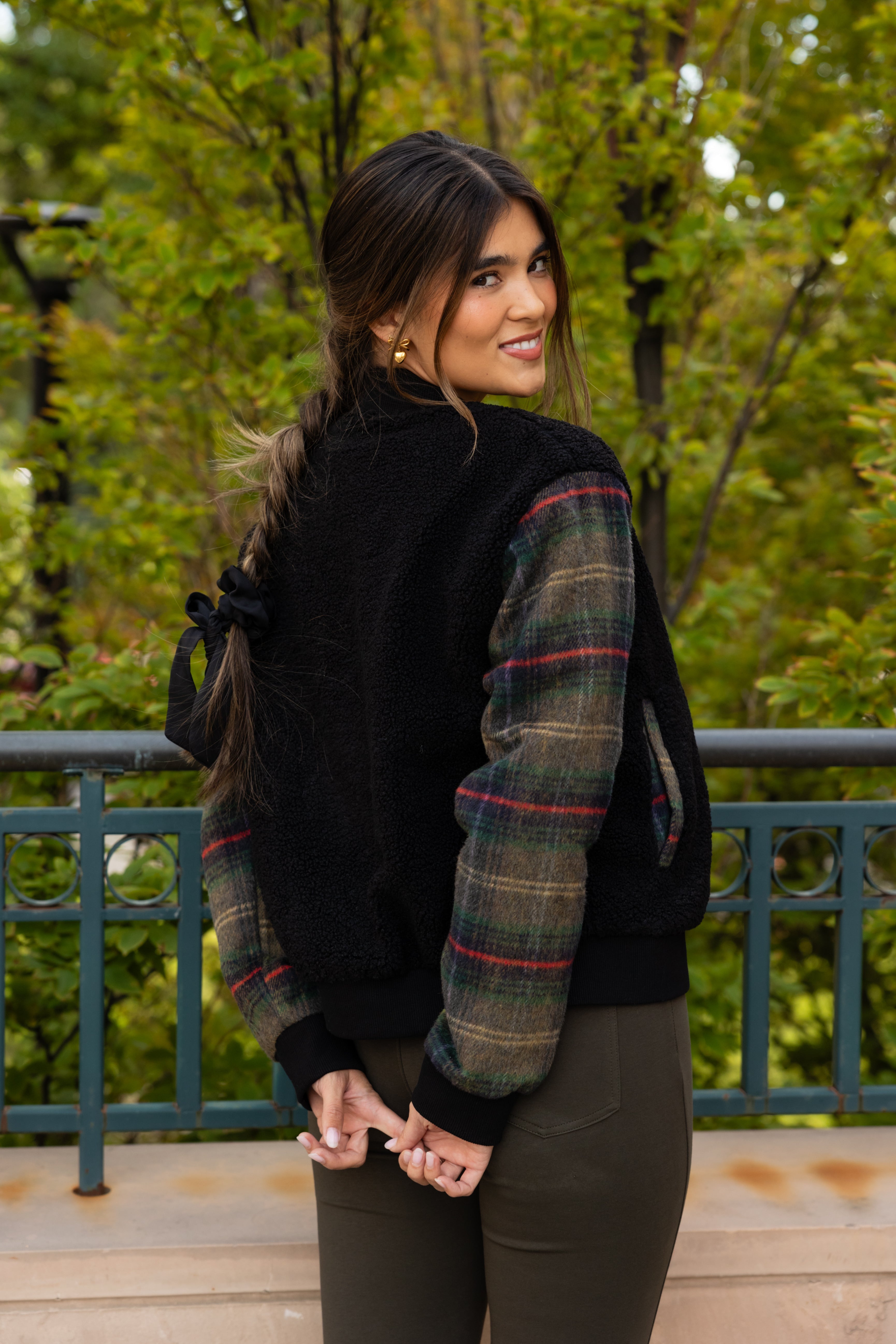A woman wearing a black fleece bomber jacket with plaid sleeves in green, red, and neutral colors, paired with dark pants, standing by a greenery backdrop.