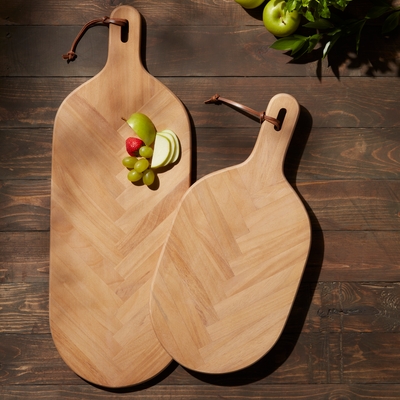 Two beech wood paddle boards with a herringbone pattern, one with a faux leather accent loop, displayed on a wooden surface with fruits and vegetables on top of one of the boards.